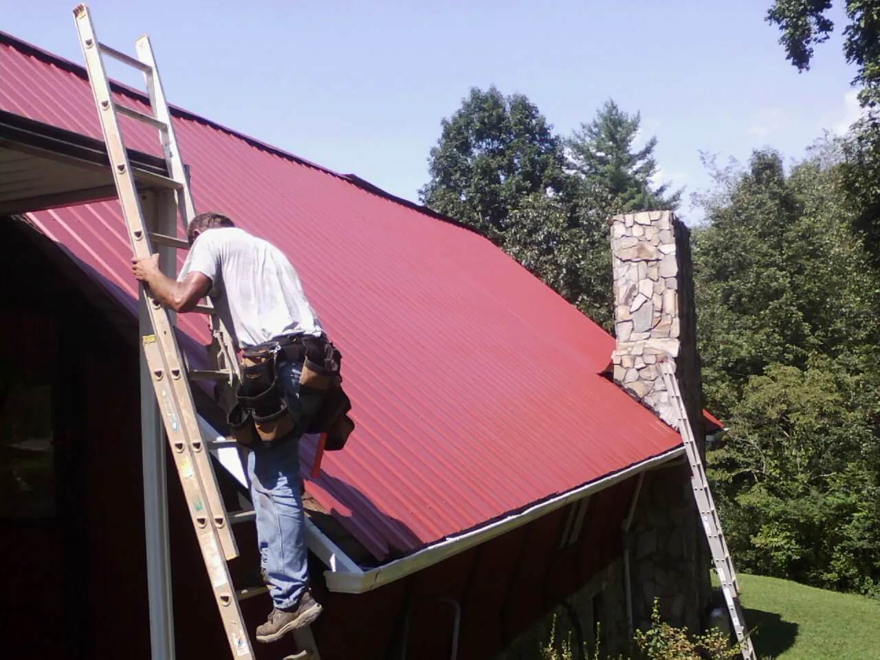 Roofer on a ladder beside a red metal roof and stone chimney — Soffit & Fascia Repair by Roofing Brothers in Radcliffe